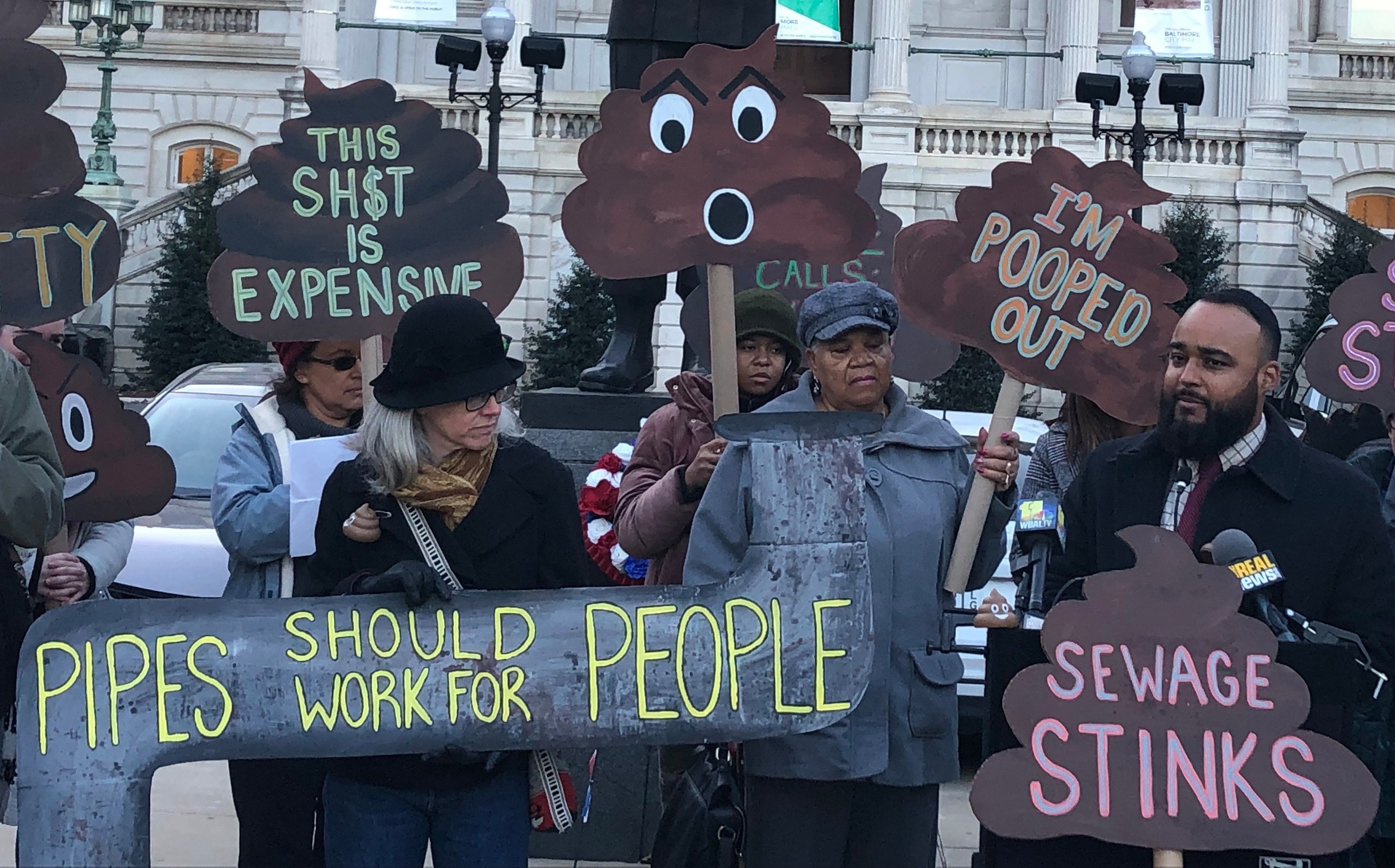 A picture of people rallying in front of Baltimore City Hall with Councilman Kristerfer Burnett speaking. People are dressed warmly and holding signs with messages like "Sewage Stinks," "I'm Pooped Out," "Pipes should work for People."